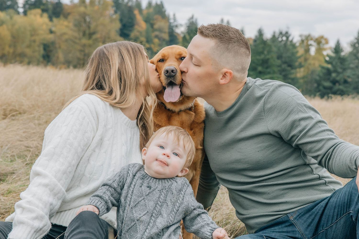 Happy couple kissing their golden retriever dog while toddler sits in front outdoors.
