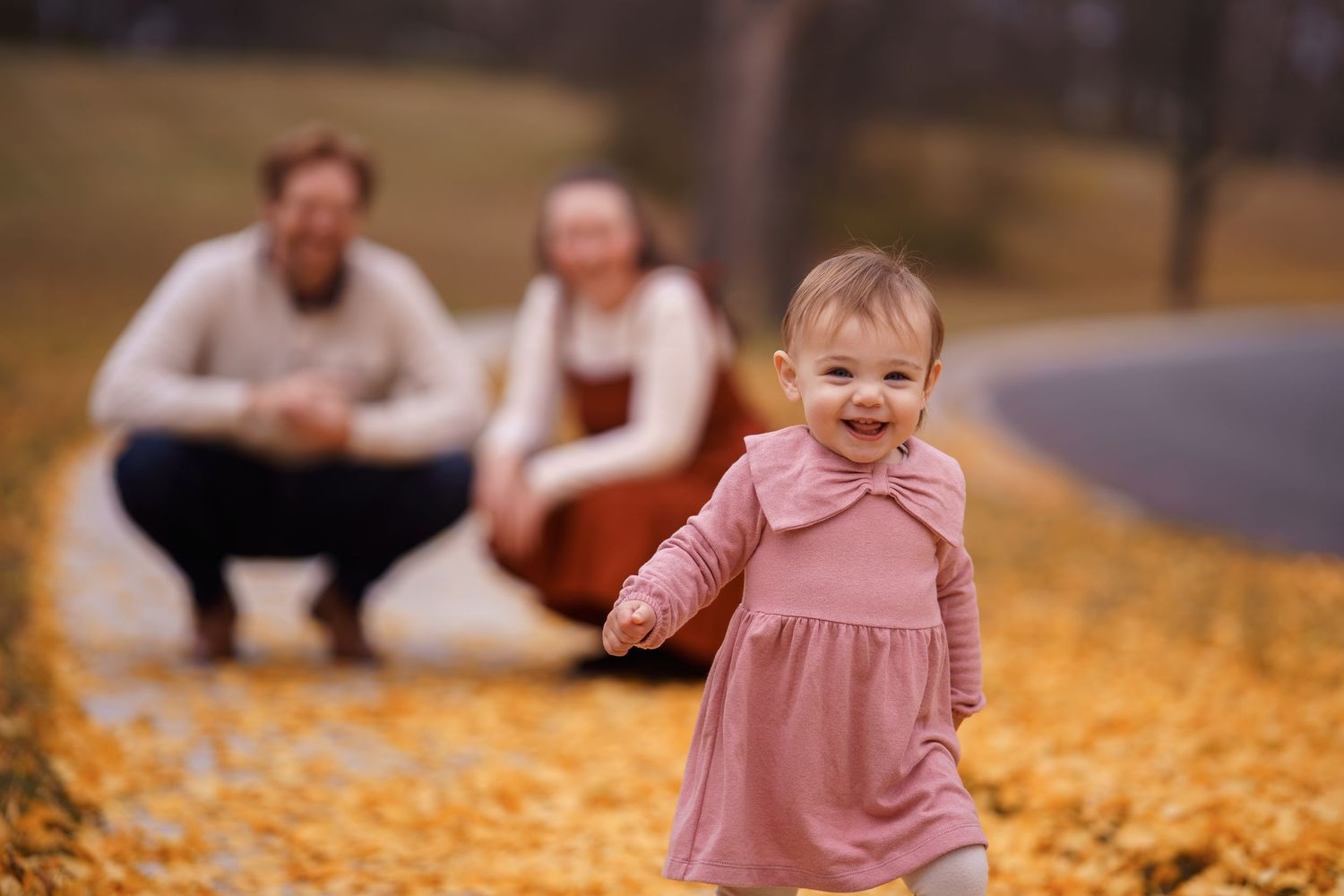 Toddler girl in pink dress running on autumn leaf-covered path with parents crouching in blurred background, in Rockville, MD