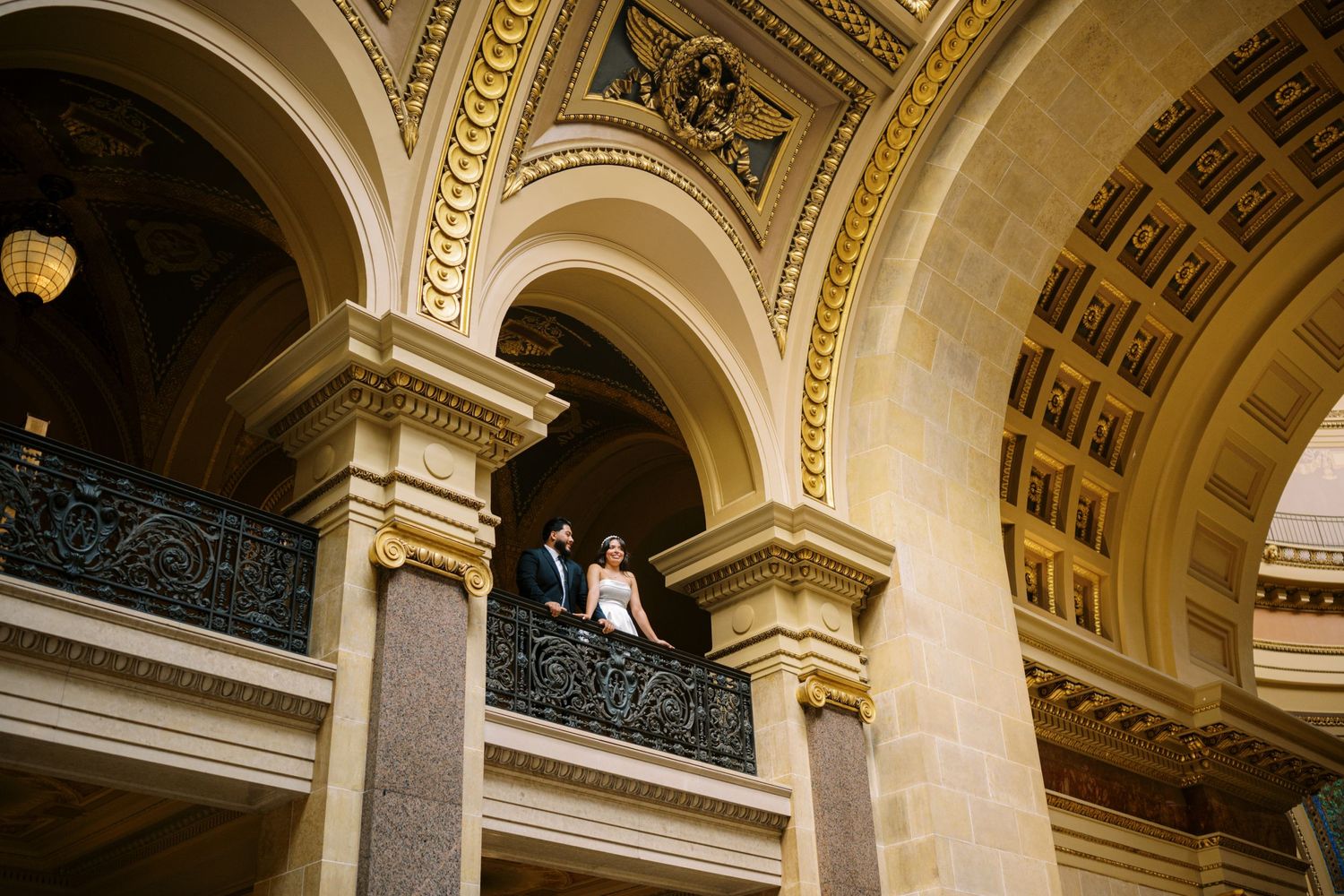 Elegant wedding couple stands between ornate marble columns and arches in a grand historic building interior.