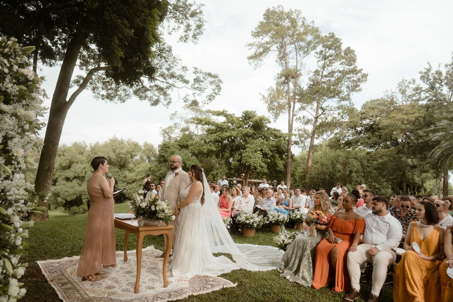 Outdoor garden wedding ceremony with guests seated on wooden chairs among tall trees.