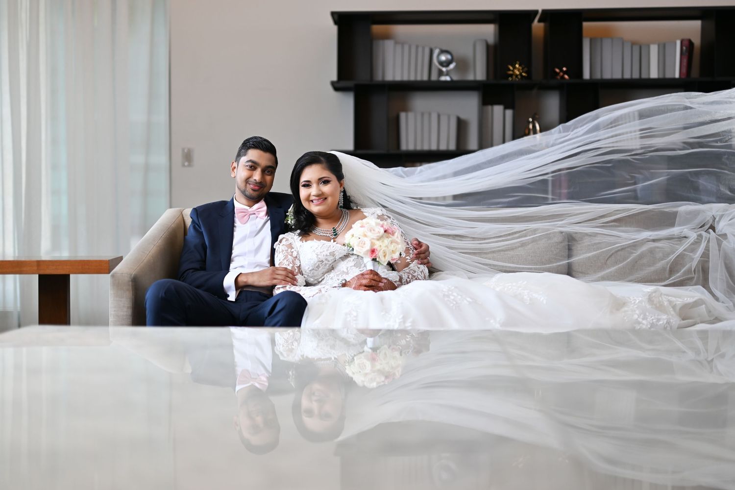 A wedding couple sits together behind flowing white veil in an elegant indoor setting.