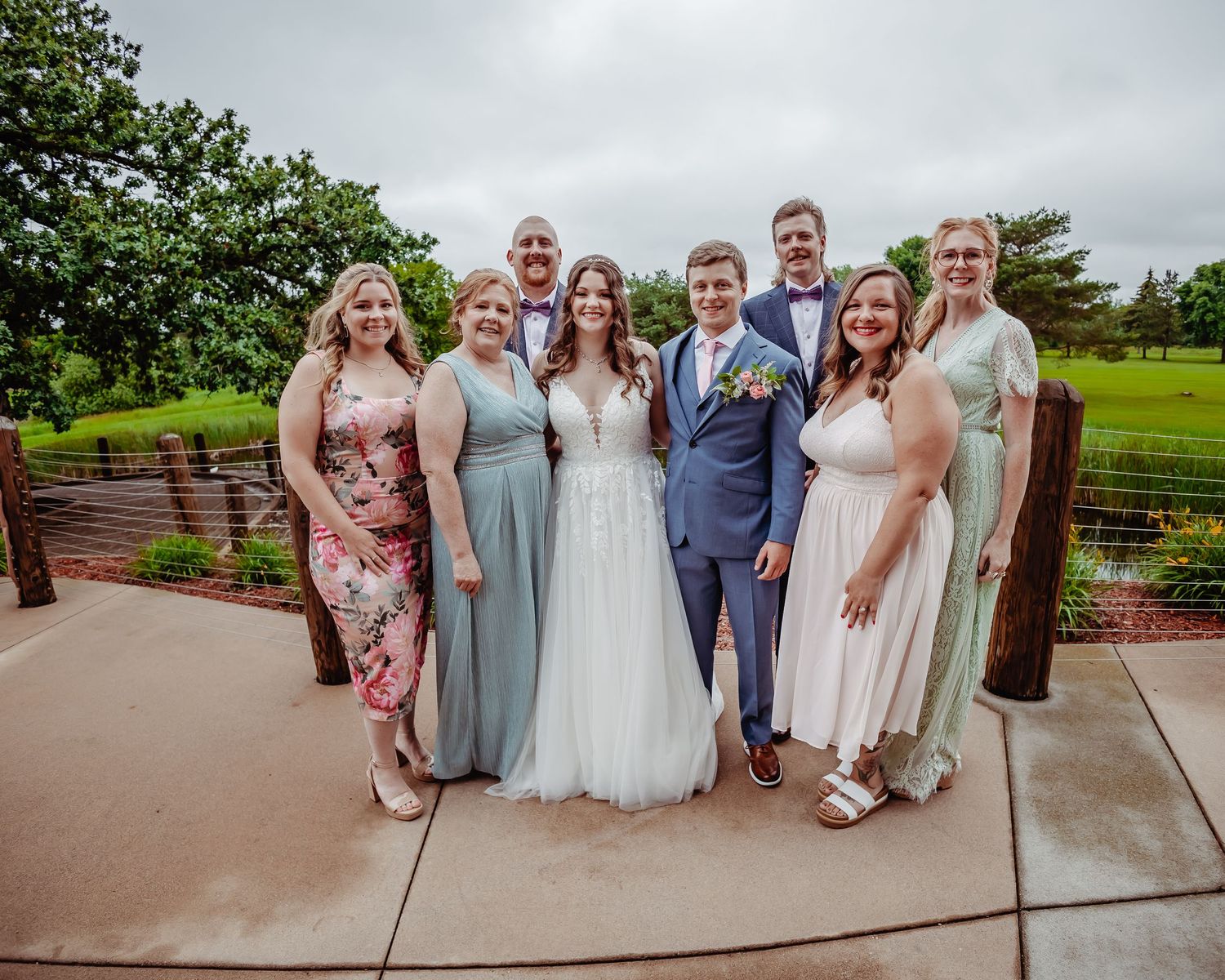 Wedding party group photo with bridesmaids and groomsmen in pastel dresses on outdoor patio.