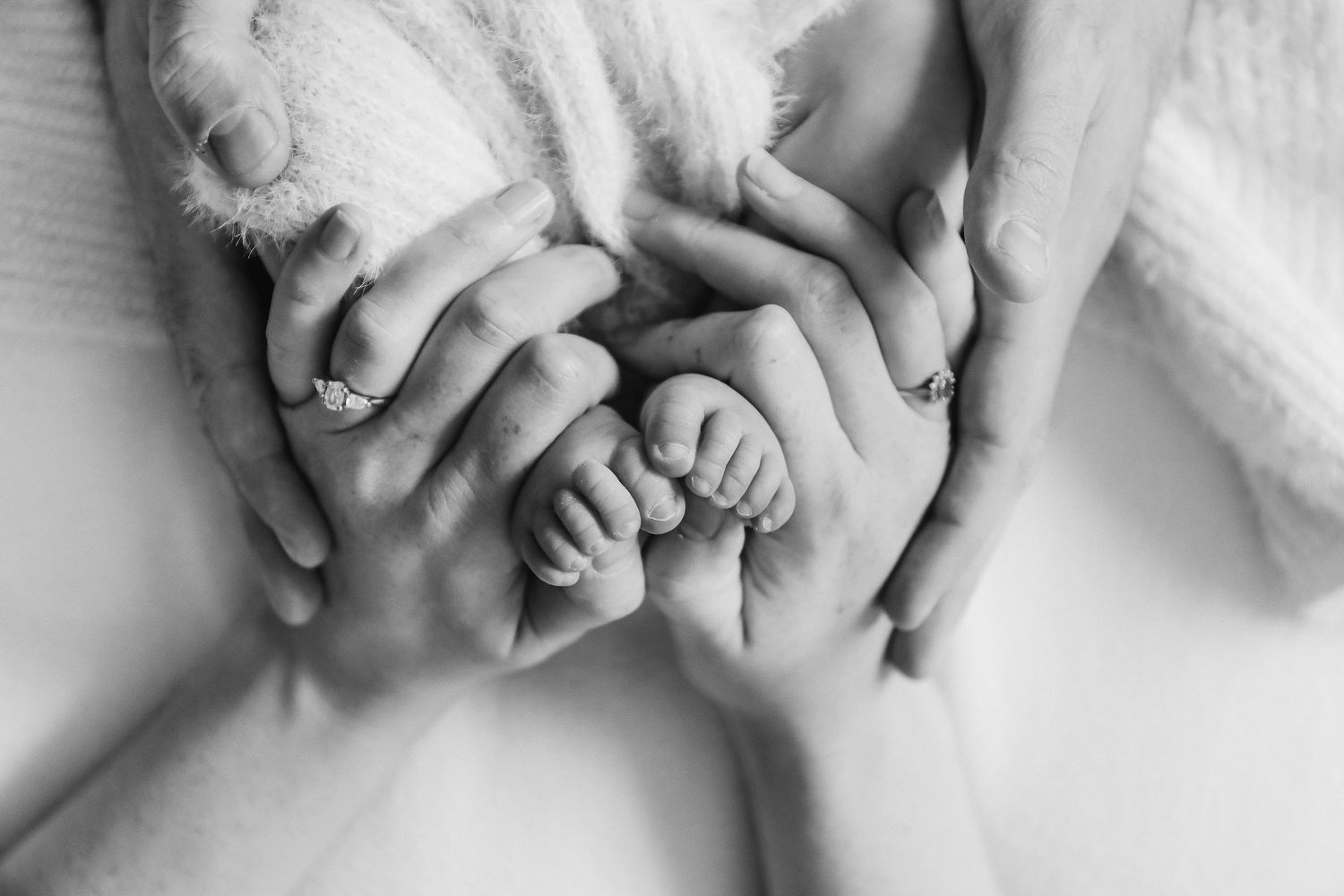 Black and white close-up of adult hands gently holding tiny baby feet wrapped in soft blankets during newborn shoot in Nottingham