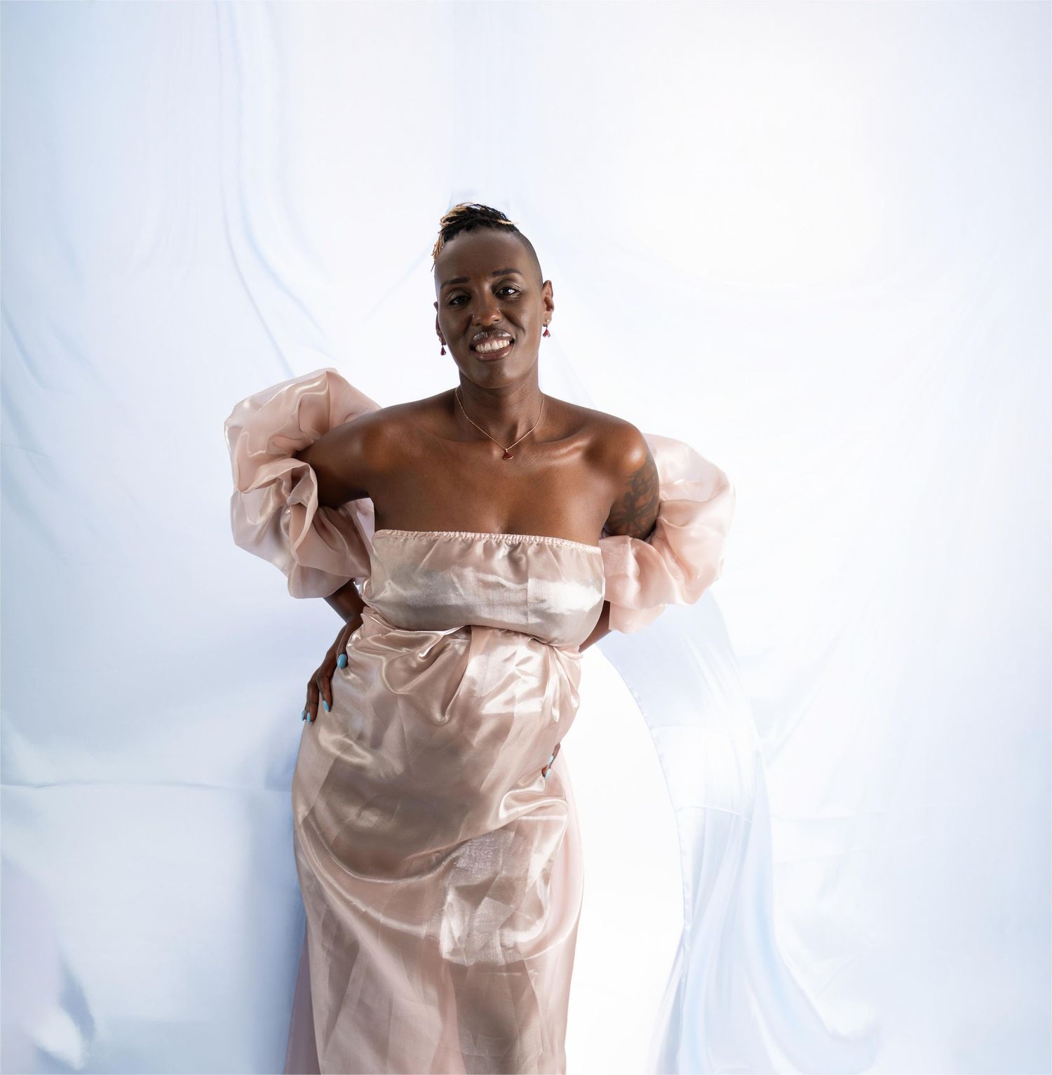 A person wearing a flowing off-shoulder light pink dress poses against a white background with ethereal fabric draping around.