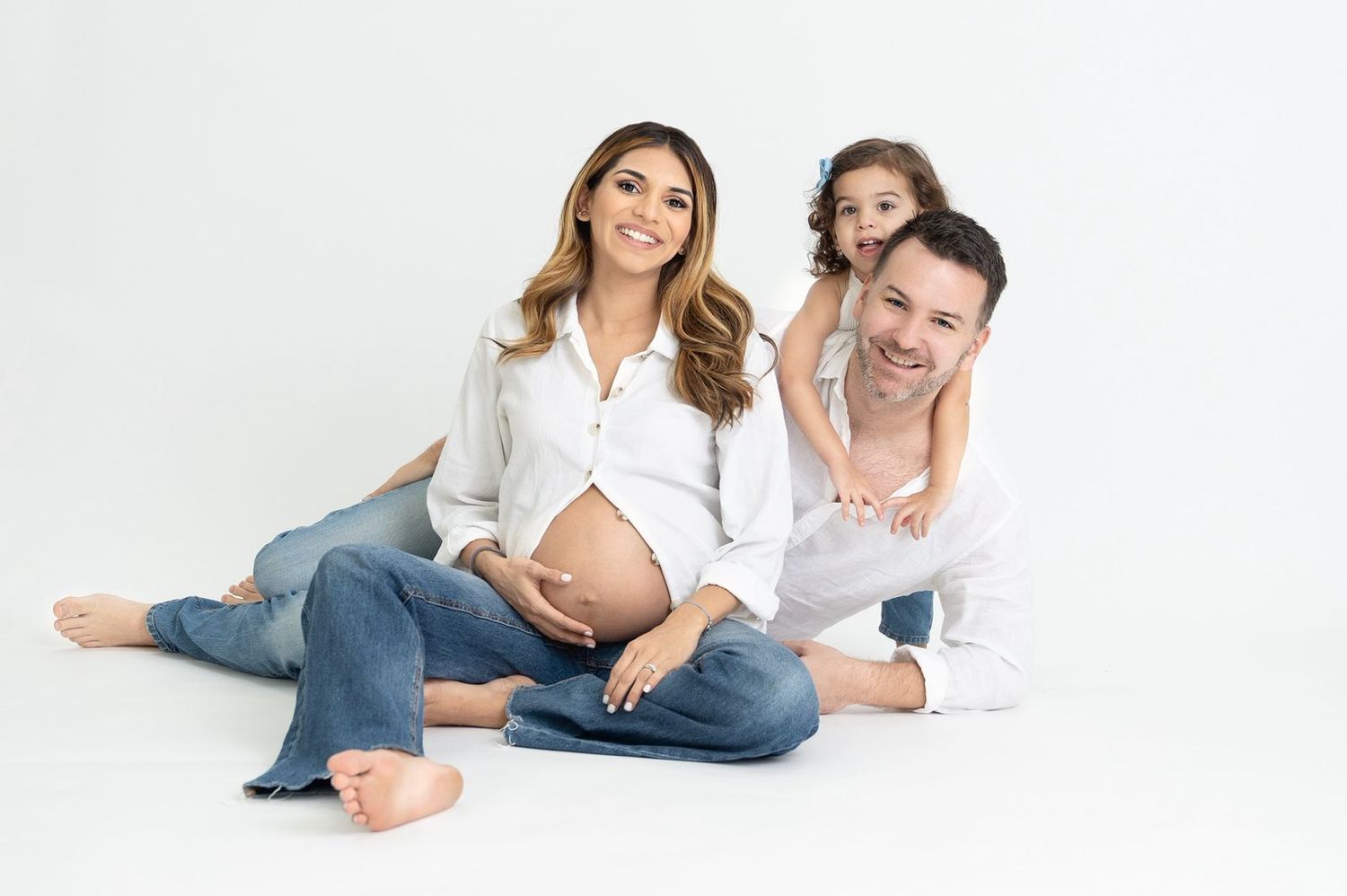 Family maternity photo with mom-to-be in white shirt and jeans posed on white background with partner and young child.