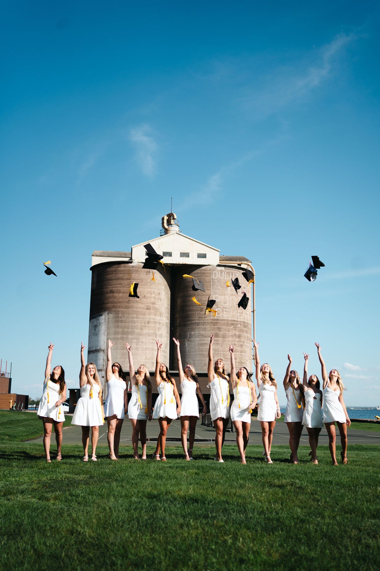 A group celebrating by tossing graduation caps in front of cylindrical silos under a blue sky.
