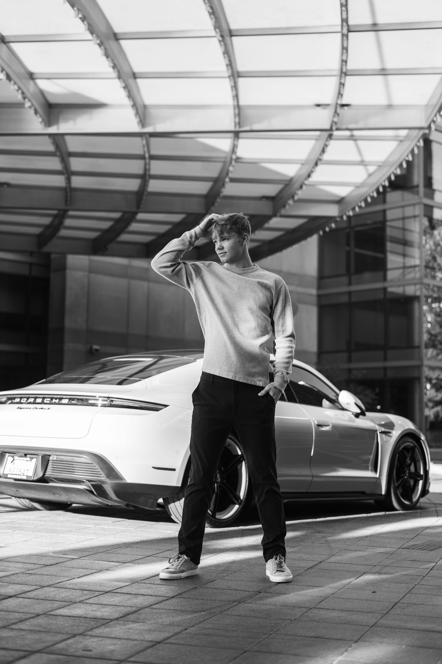 Black and white photo of someone posing in casual wear next to a luxury sports car under a covered outdoor area.