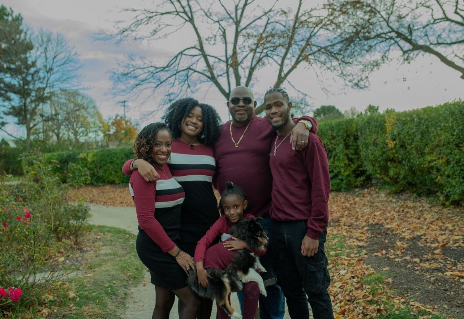 A group in coordinating burgundy outfits poses together on an autumn trail with bare trees in the background.