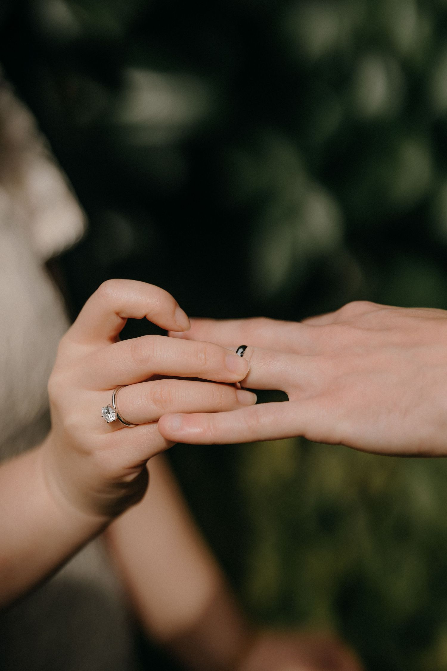 Close-up of hands exchanging wedding rings during an intimate outdoor ceremony with blurred green foliage background.
