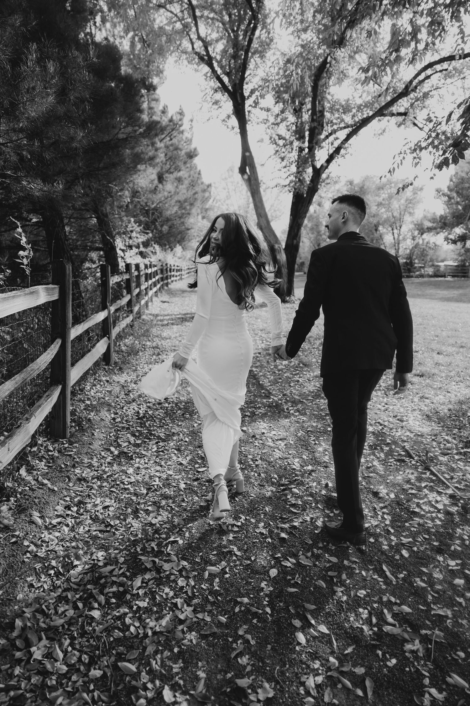 Black and white photo of bride and groom holding hands walking along a tree-lined path.