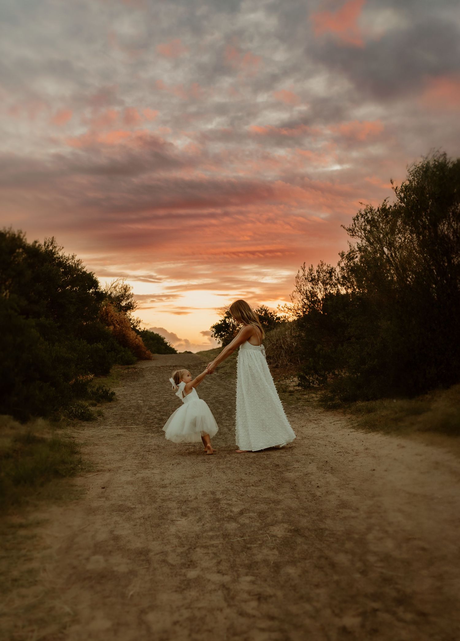 A white flowing dress catches the light of a dramatic sunset on a sandy path through coastal vegetation.