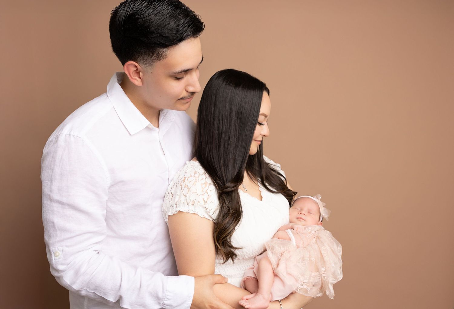 Couple in white and pink outfits embrace lovingly in a soft, romantic studio portrait with warm brown background.
