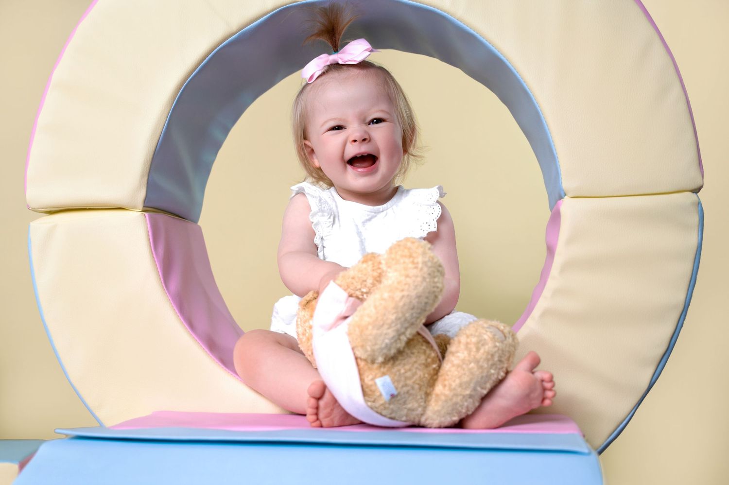 A laughing baby in white clothing sits inside a pastel-colored play tunnel while holding a teddy bear.