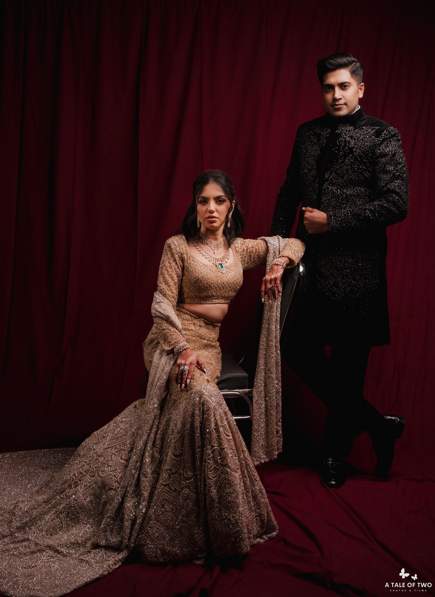 A couple in formal attire poses dramatically against a dark red backdrop in a romantic photoshoot setting.