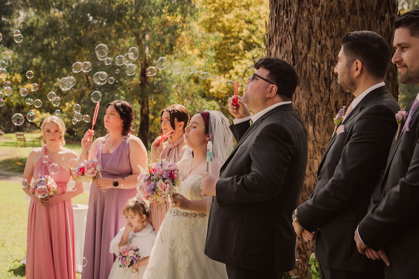 Wedding party poses together in an outdoor garden setting during autumn with golden foliage in the background.