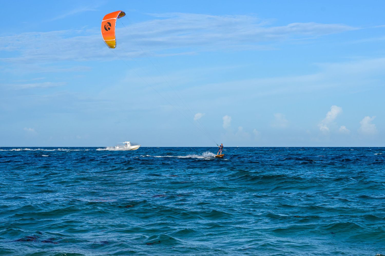 Kitesurfer riding ocean waves with an orange kite against a blue sky with a boat in the background.