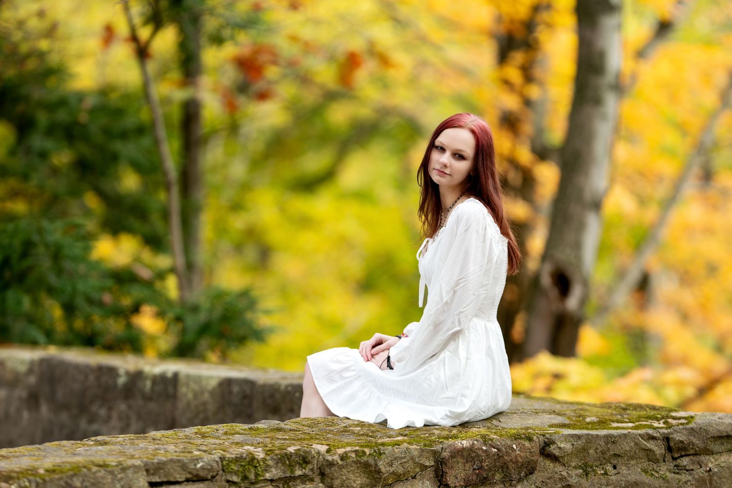 Young redhead woman in white dress sitting on stone wall in autumn park.