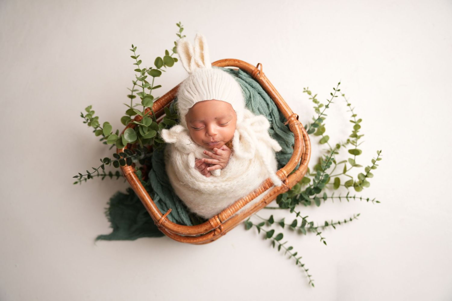 A newborn in a white wrap sleeps peacefully in a wooden basket surrounded by green eucalyptus sprigs.