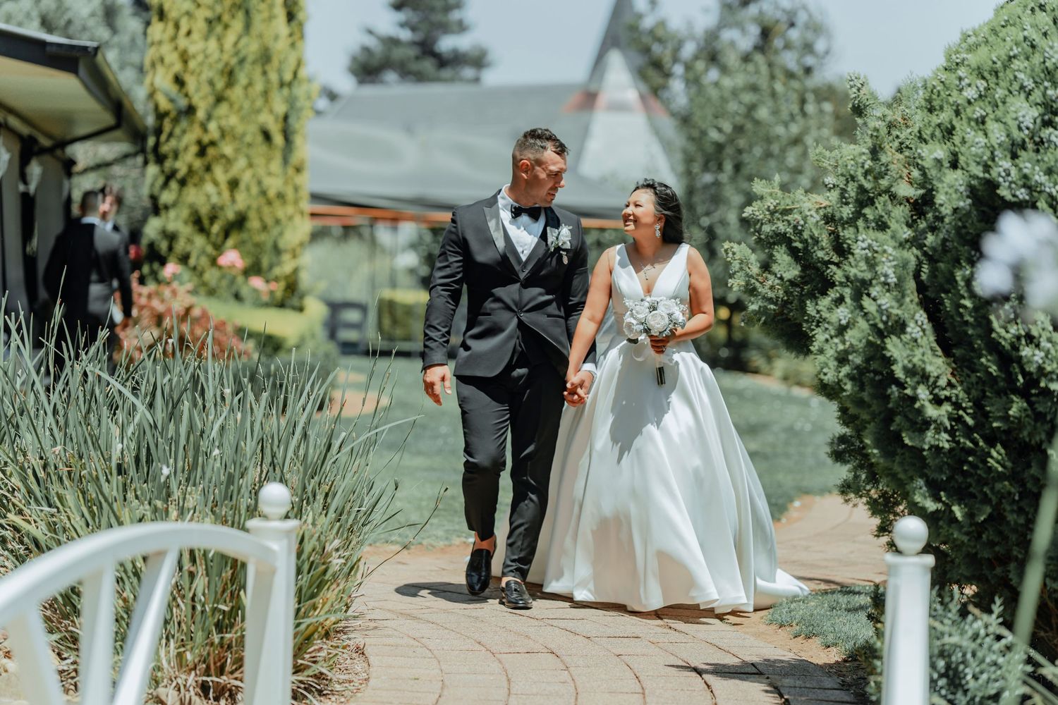 Wedding party walks along a garden path during a sunny outdoor celebration at an elegant venue with lush greenery.