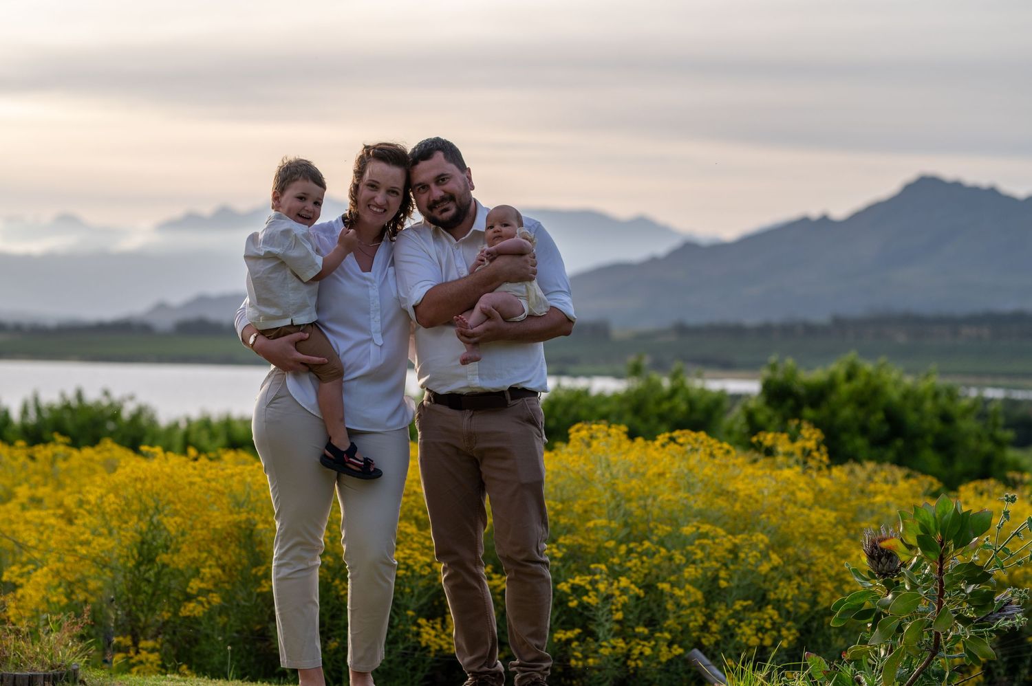 Family poses together in a golden yellow wildflower field at sunset with mountains in the background.