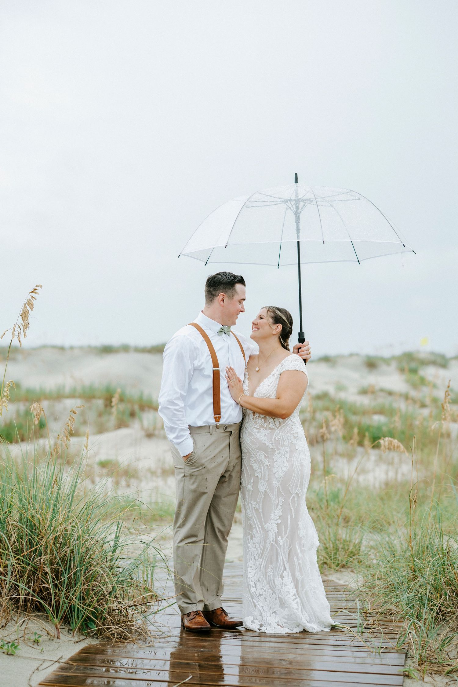 A romantic couple poses with a white umbrella on a wooden boardwalk along sand dunes at the beach.