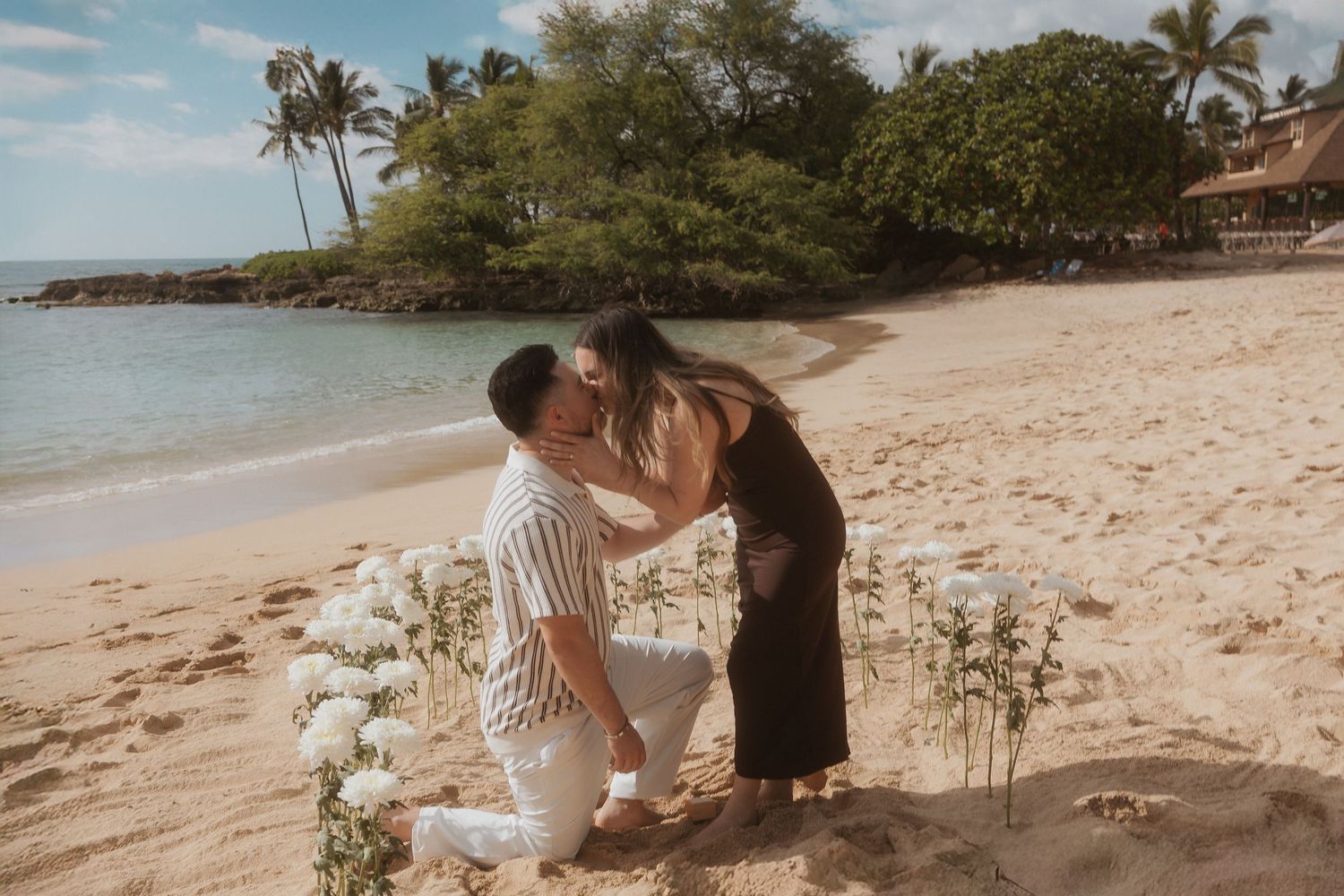 A romantic beach proposal scene with candles and flowers arranged on a tropical Hawaiian shoreline with palm trees at sunset.