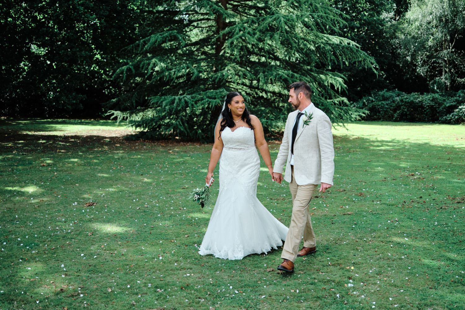A couple walks hand in hand across a sunlit lawn surrounded by lush green trees during their outdoor summer wedding at Oakwood House