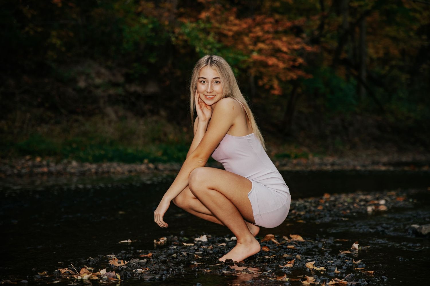 Young person in white tank top poses playfully against bokeh background with natural lighting.