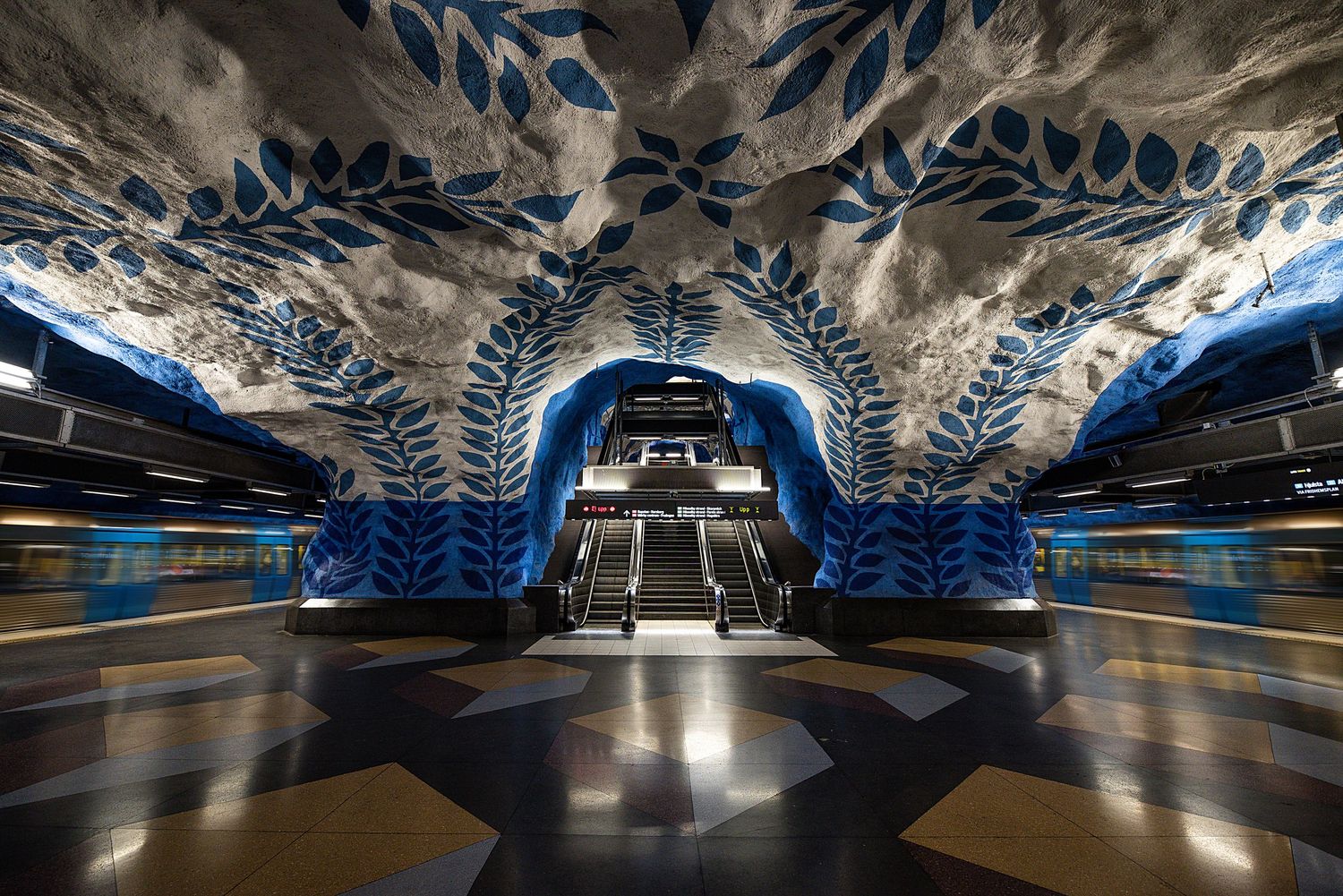Futuristic subway station interior with ornate blue floral patterns and symmetrical lighting design.
