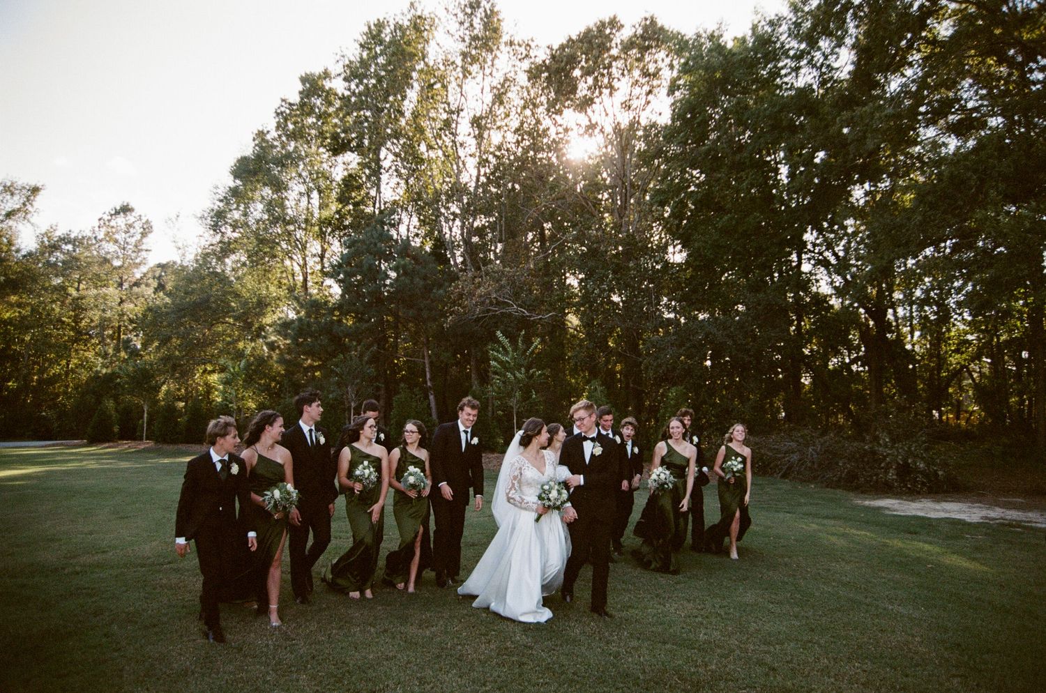 Wedding party poses together on a grassy hill during golden hour sunset at an outdoor celebration.