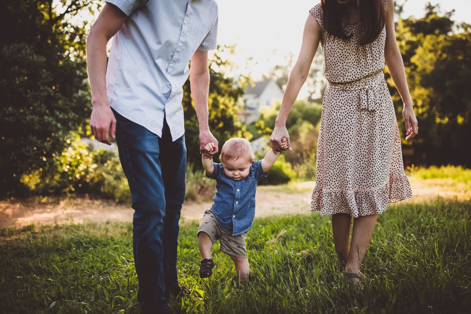 A couple and their toddler walk together through a sunlit grassy field during golden hour.