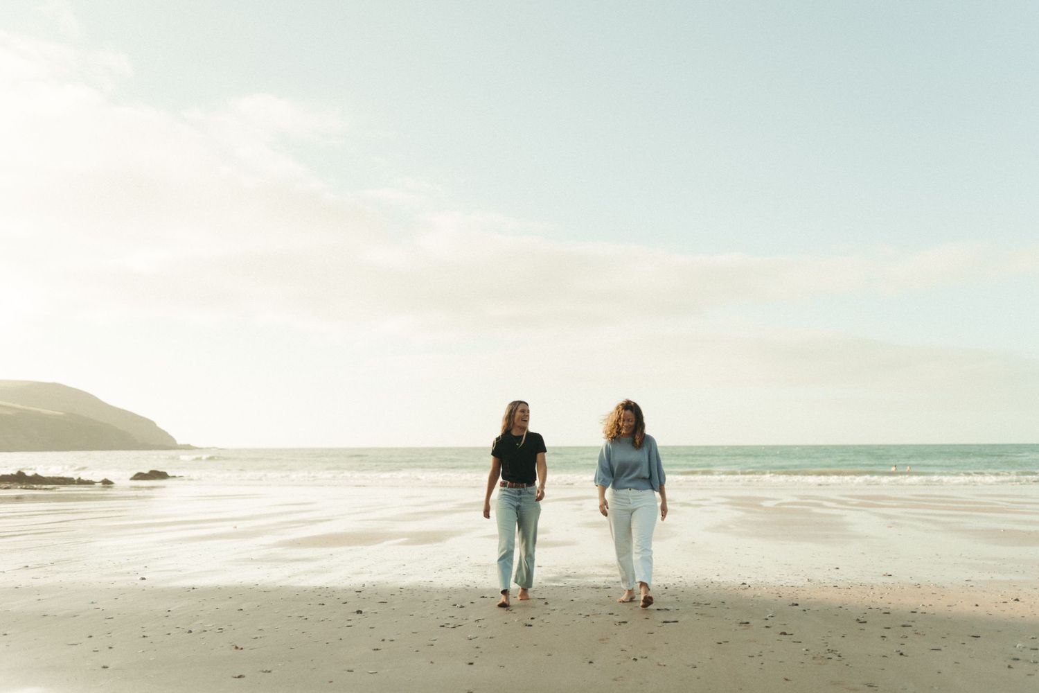 Two people walk along a misty beach shoreline with rolling waves and mountains in the background.