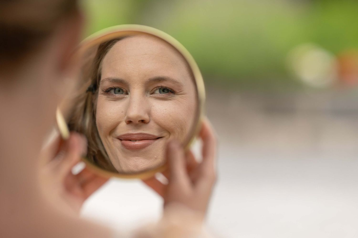 A person's reflection is seen smiling in a handheld mirror against a soft green blurred background.