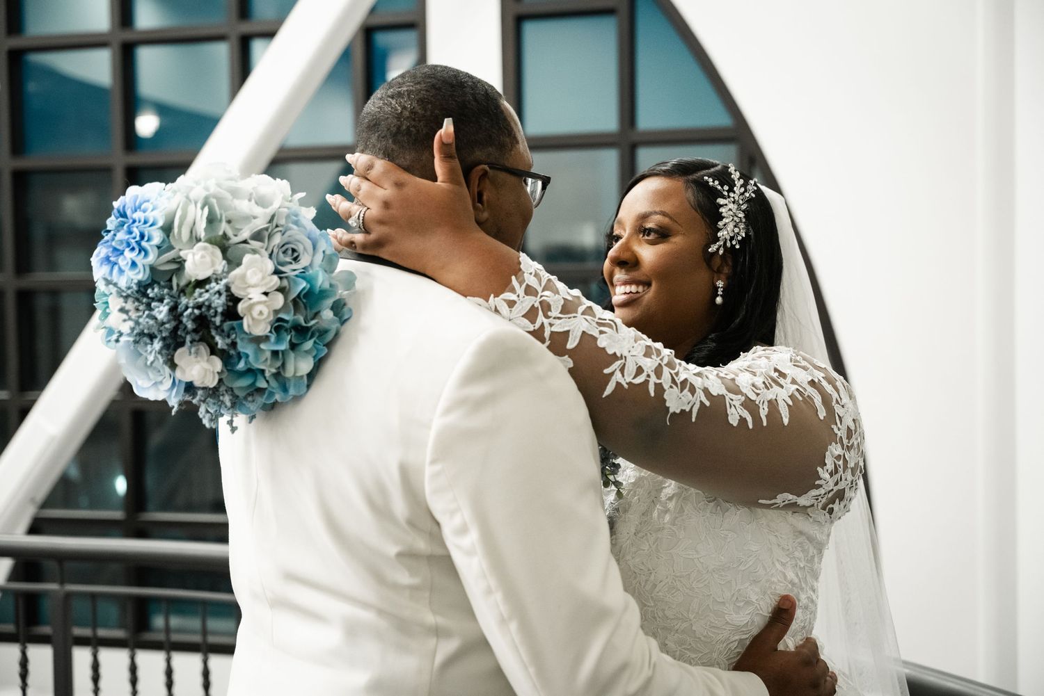 A joyful bride in a lace wedding gown with long sleeves holds a blue bouquet while celebrating with her groom in white.