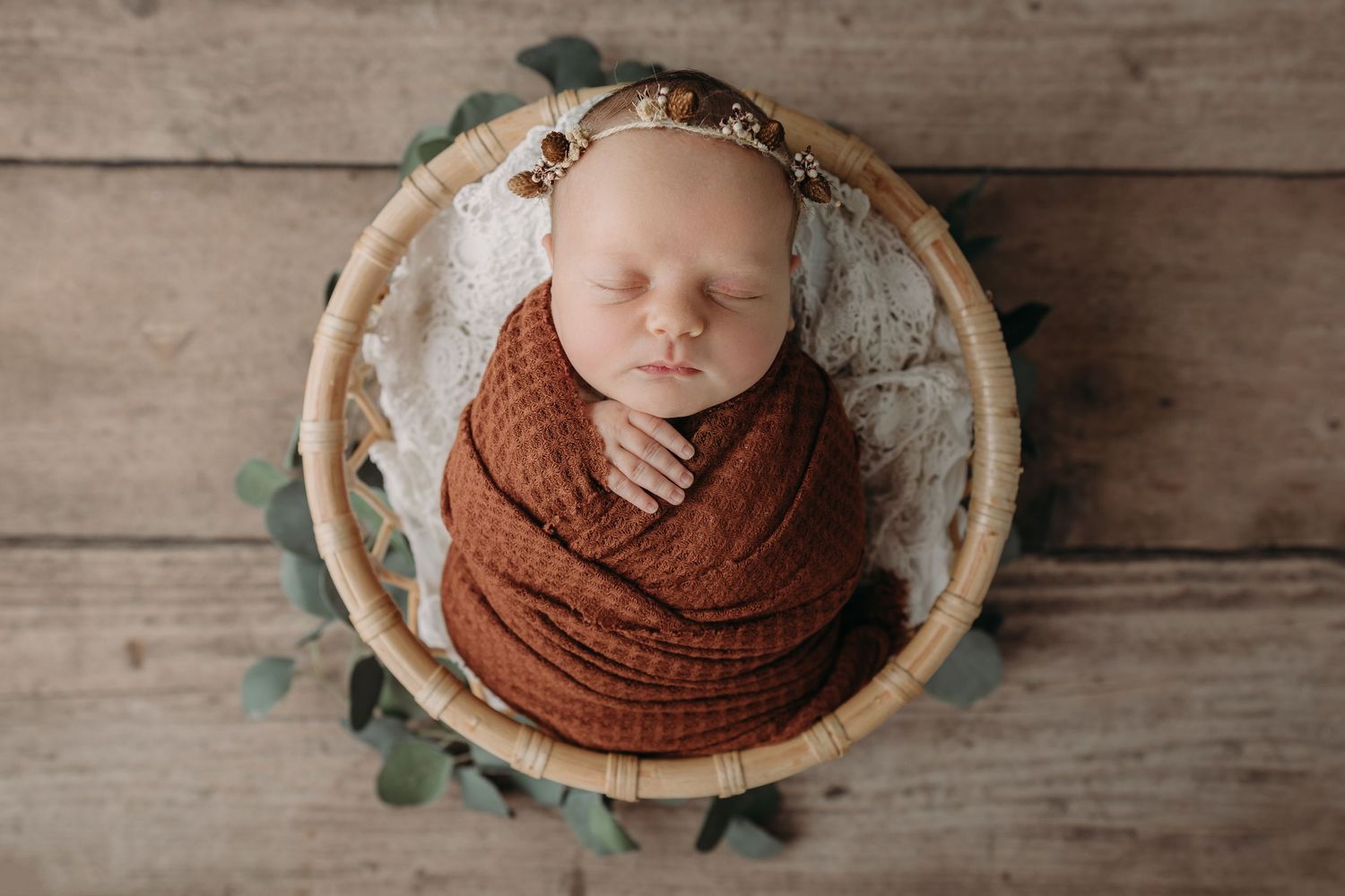 A sleeping newborn wrapped in a rust-colored knit blanket rests in a wooden bowl with greenery accents.
