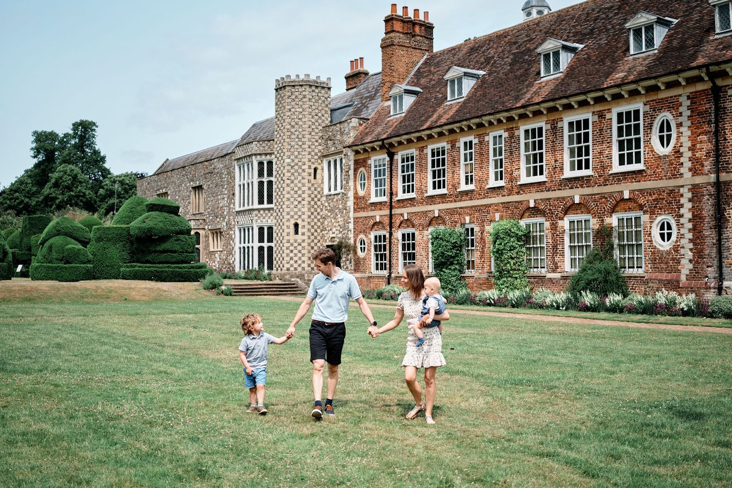 People walk on a sprawling lawn in front of a historic English manor house with towering chimneys and classic Georgian windows.