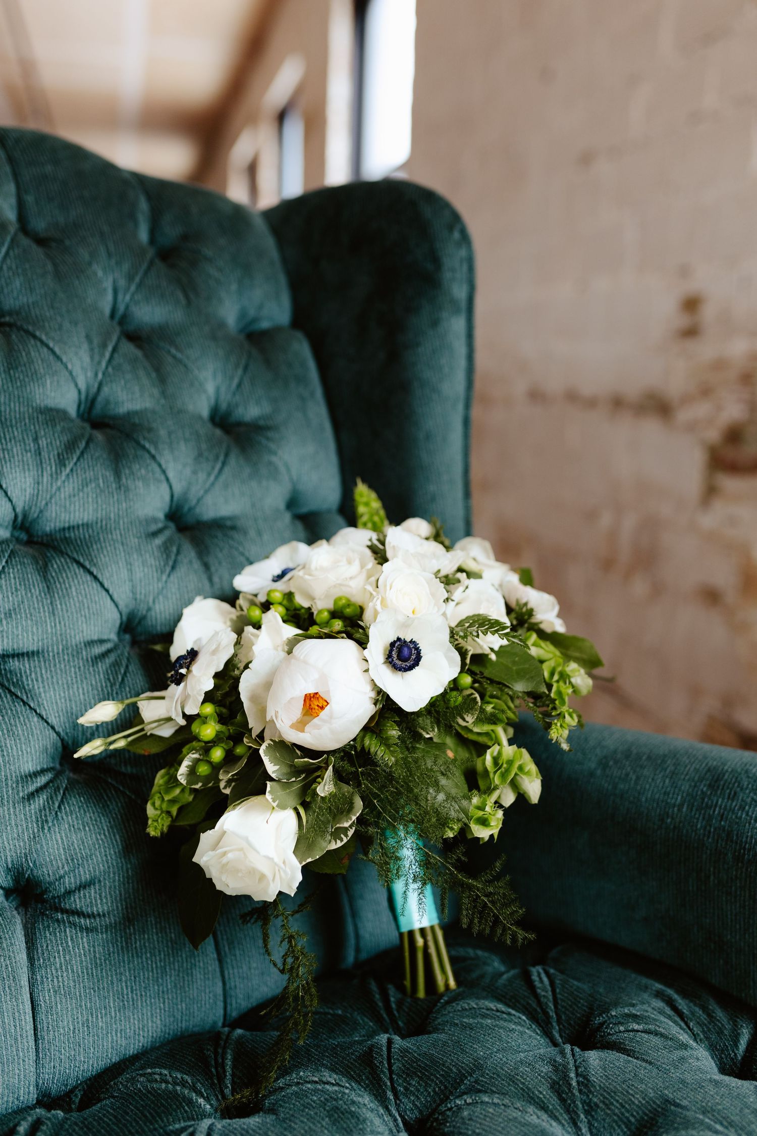 Wedding bouquet with white anemone flowers and greenery resting on emerald green tufted velvet chair.