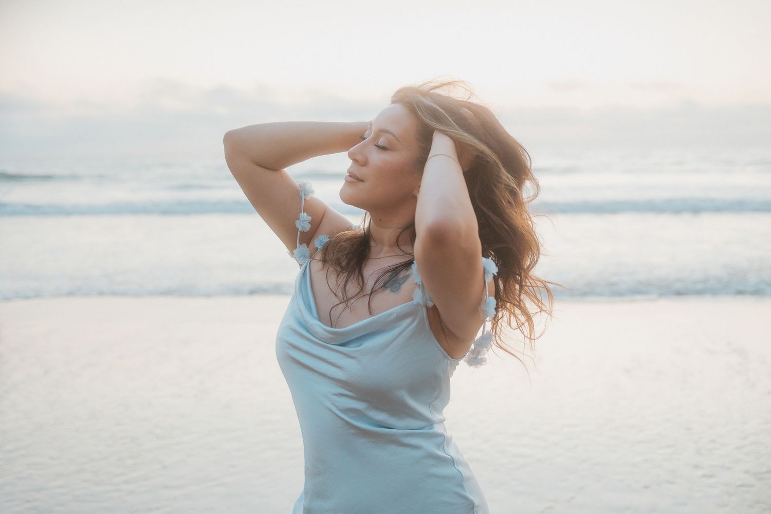 Woman in flowing blue dress dances gracefully on beach at sunset with hair blowing in ocean breeze.