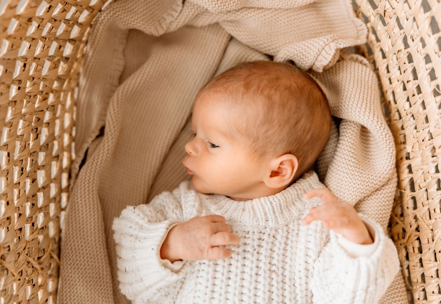 Baby wearing white knit sweater rests in woven bassinet with beige bedding.