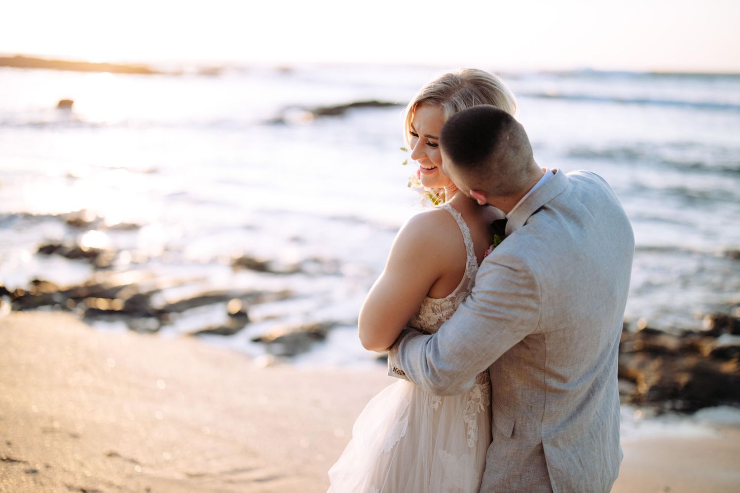 Couple embraces on a rocky beach at sunset with ocean waves crashing in the background.