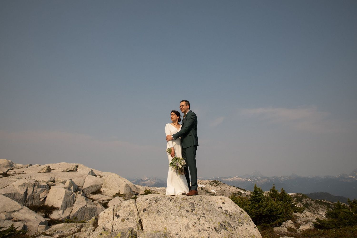 Bride and groom stand together on rocky mountain summit under clear blue sky during outdoor wedding.
