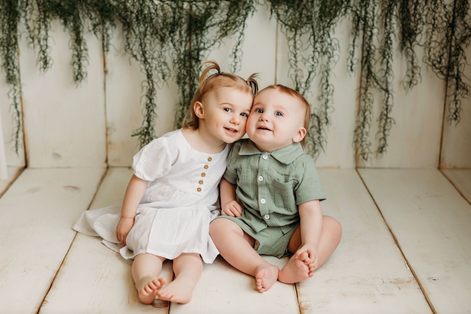 Two small children sitting close together in white photo backdrop with hanging greenery decor above.
