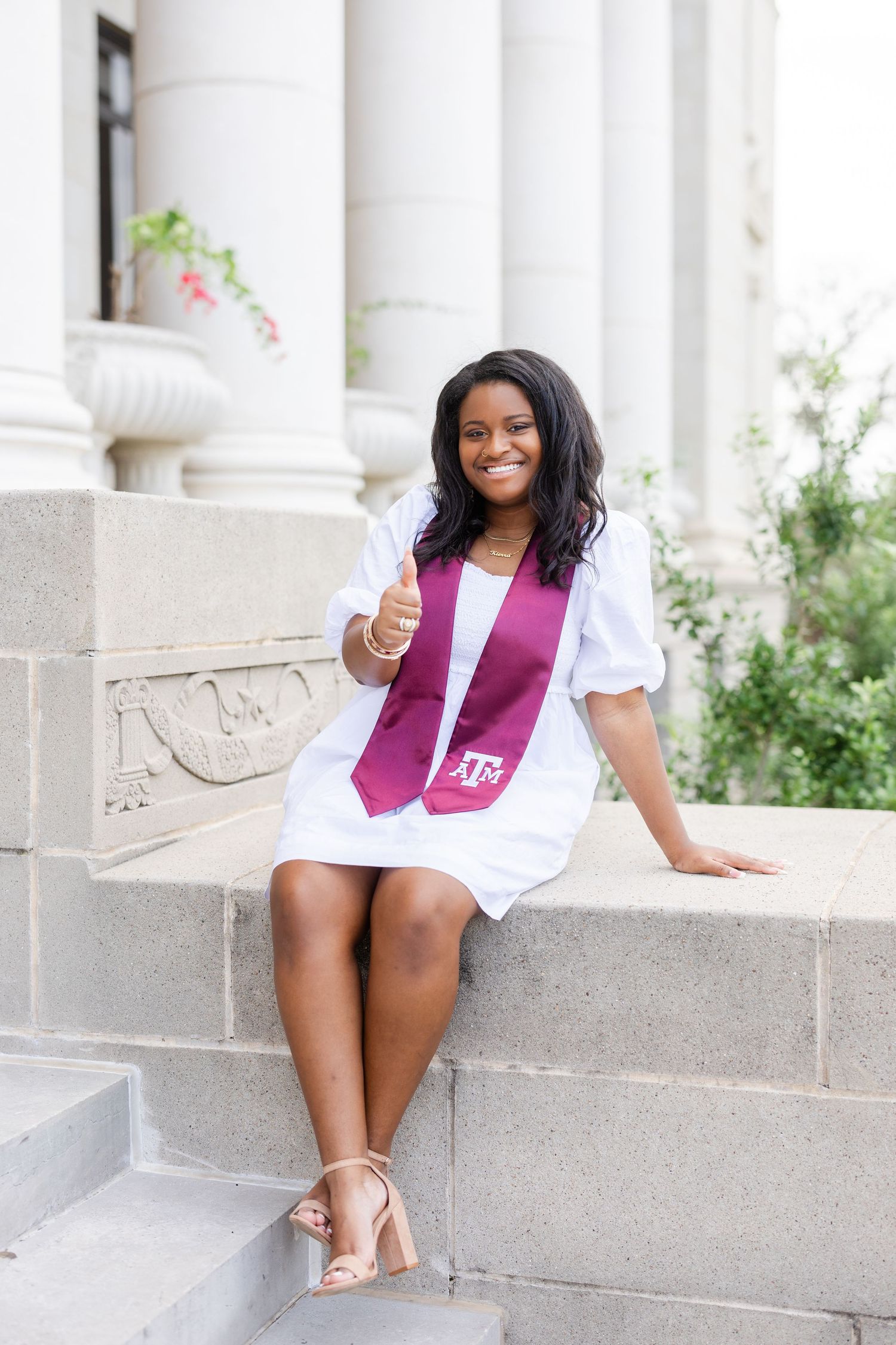 Graduate in white dress and maroon stole sits on stone steps near columns.