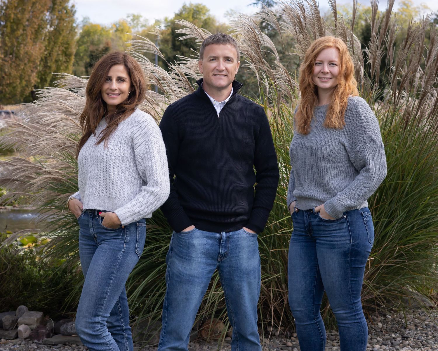 Three people in casual sweaters and jeans pose together against tall ornamental grasses.