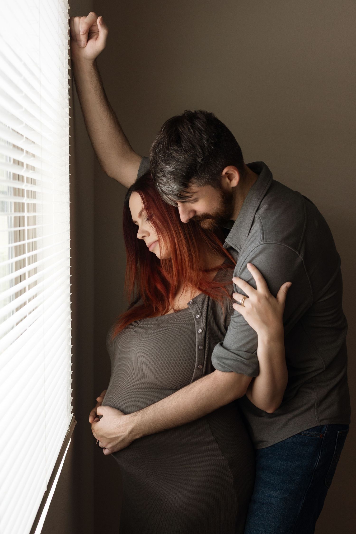 A bearded man tenderly embraces his pregnant redhead partner near a window with blinds in a cozy indoor setting.