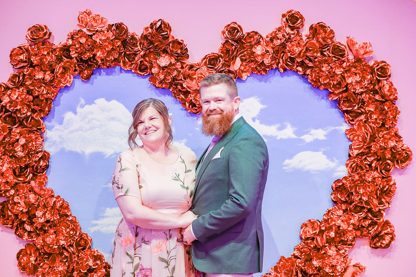 A romantic couple poses under a series of red rose heart-shaped arches against a pink and blue sky background.