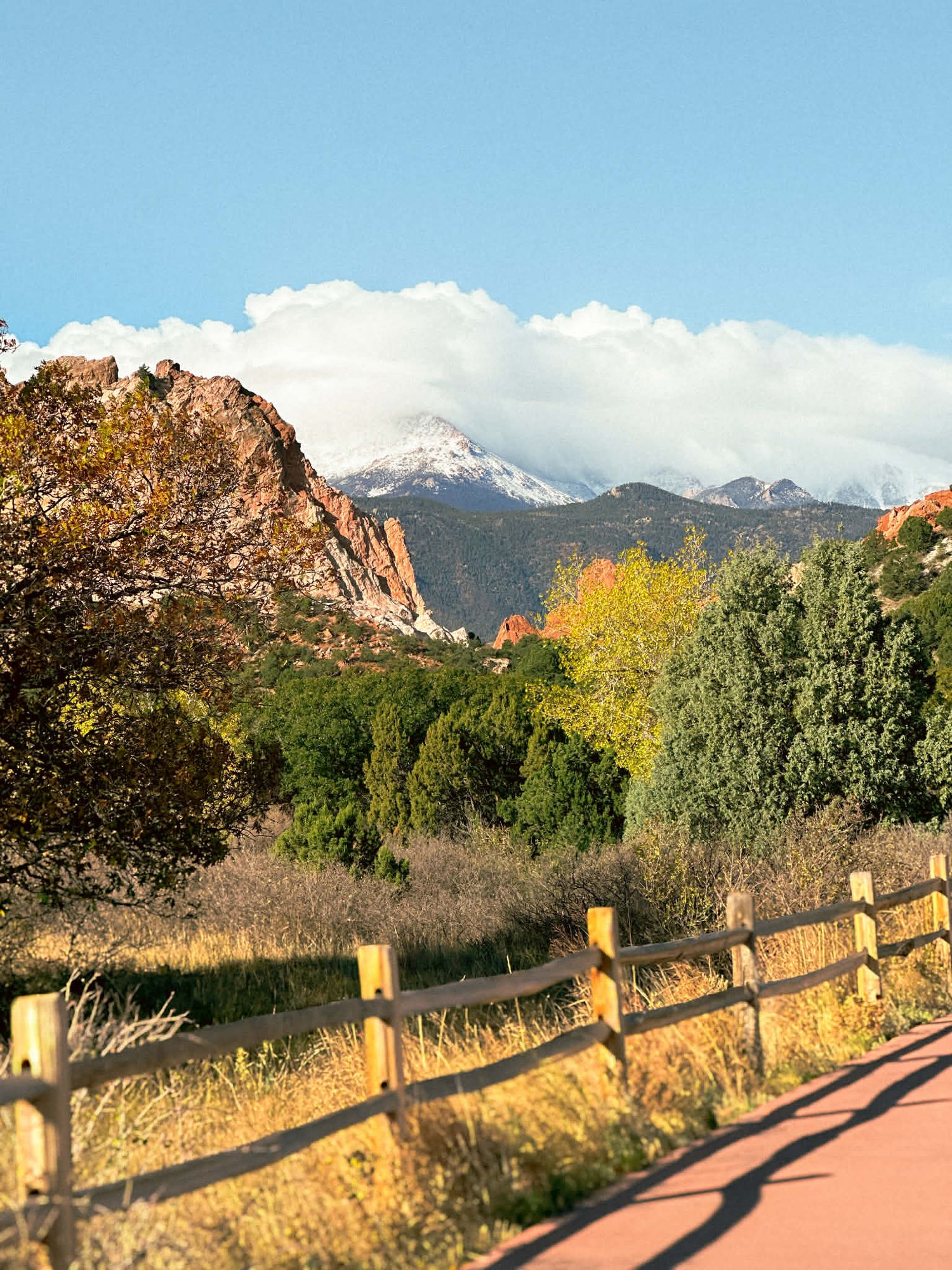 A scenic trail path with wooden fencing leads through Garden of the Gods park with red rock formations and mountains.