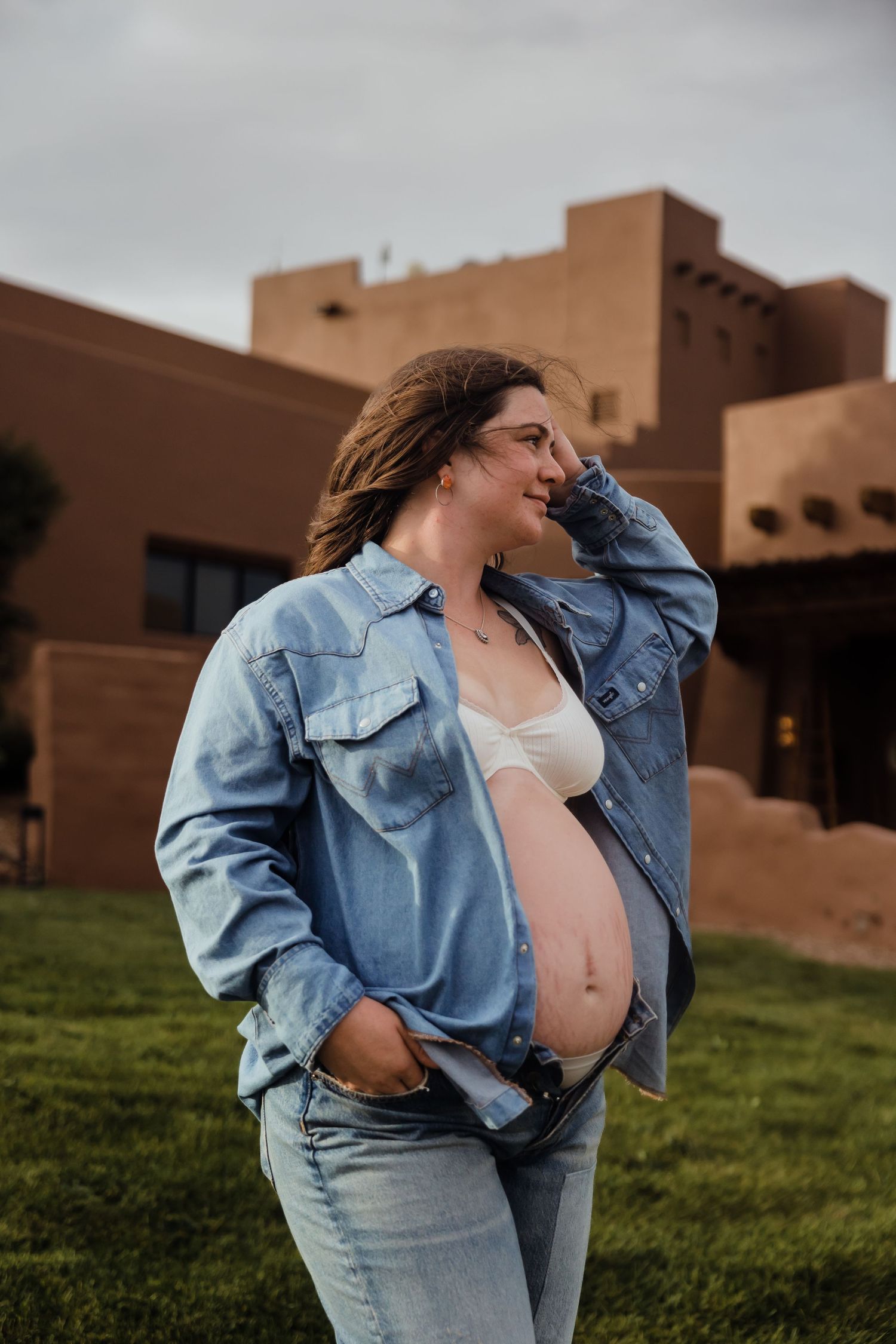 Pregnant woman in open denim shirt and white bralette poses outdoors near adobe building on cloudy day.
