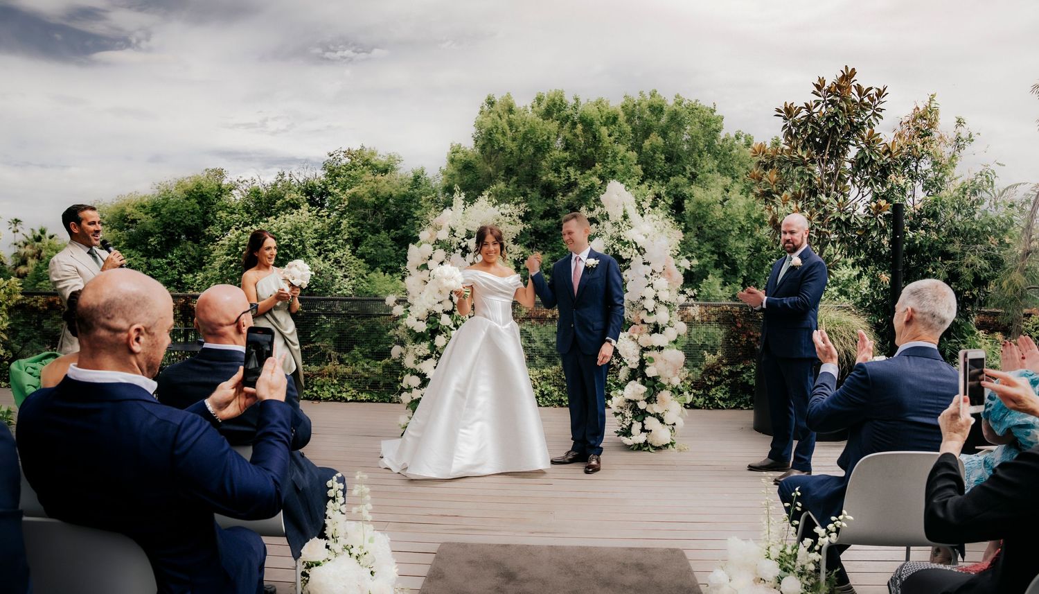 Wedding ceremony held outdoors on a patio with guests taking photos and floral arch decorations under cloudy skies.