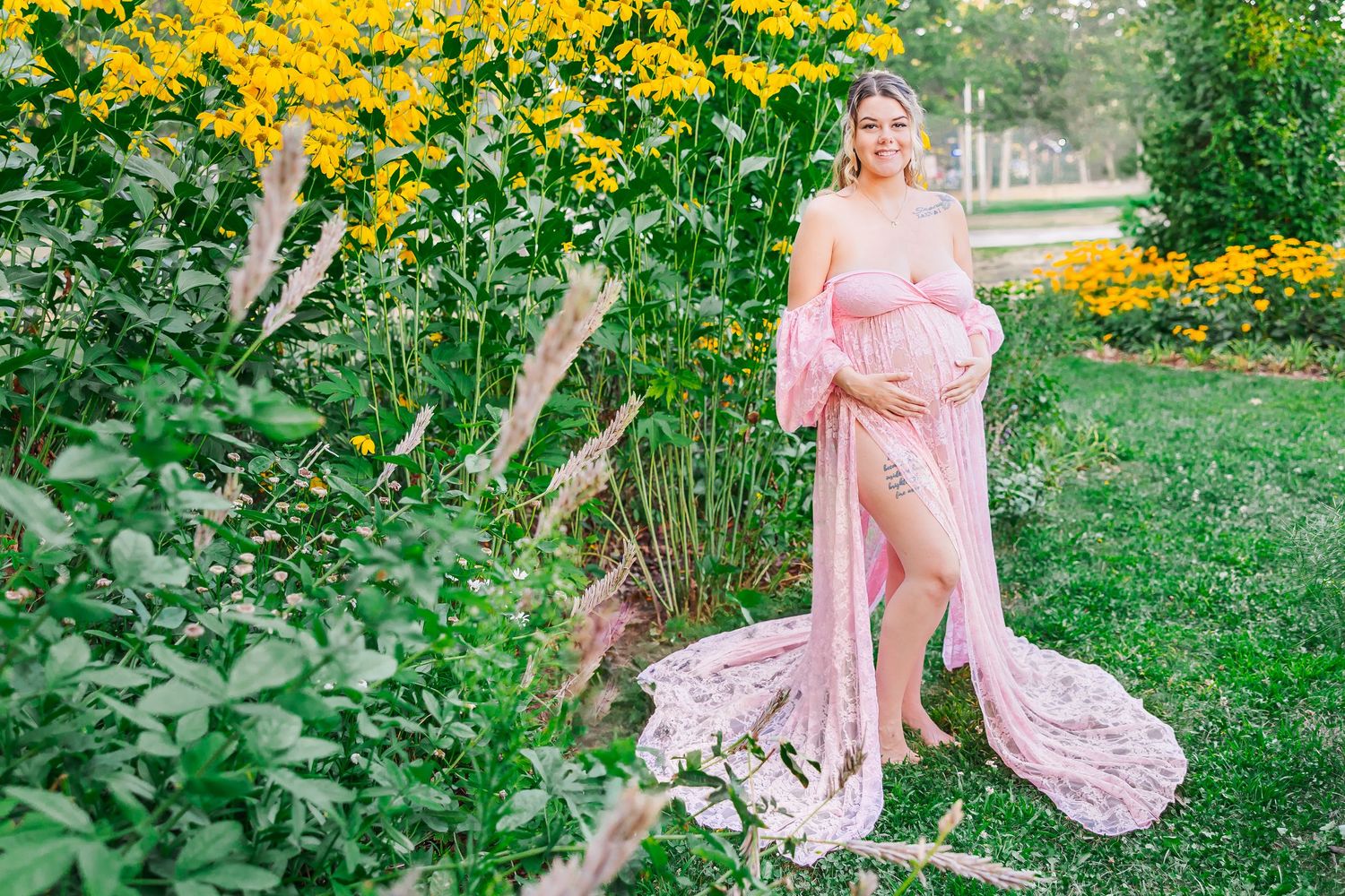 A figure in a flowing pink maternity gown poses in a garden with bright yellow flowers and greenery.